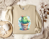 a beige sweater with a printed design of a cactus and the text "Don't be a prick" on it. The sweater is laid out on a bed with a white blanket and a white mug. There are also some white flowers and a white snowflake decoration in the scene.