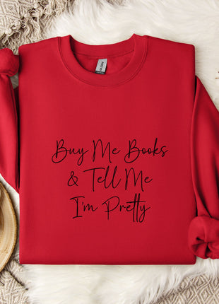 A red sweater with the text "Buy Me Books & Tell Me I'm Pretty" printed on it, along with a mug and some white flowers on a white fur rug.