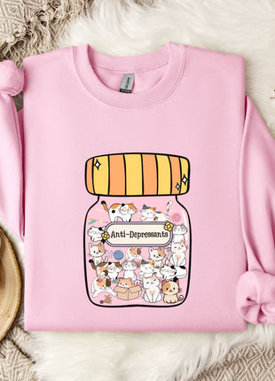 A pink sweatshirt with a graphic of a jar filled with cats and the text "Anti-Depressants" printed on it, along with a wooden tray and some white flowers.