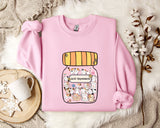 A pink sweatshirt with a graphic of a jar filled with cats and the text "Anti-Depressants" printed on it, along with a wooden tray and some white flowers.