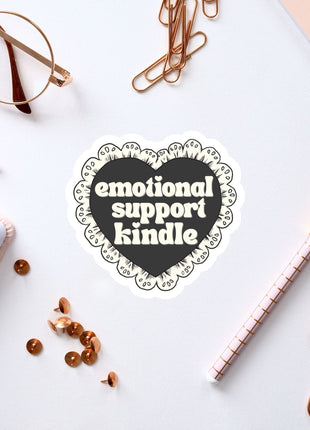a heart-shaped sticker with the text "emotional support Kindle" surrounded by various office supplies such as a pen, a notebook, and a pair of glasses.
