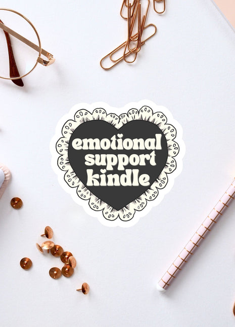 a heart-shaped sticker with the text "emotional support Kindle" surrounded by various office supplies such as a pen, a notebook, and a pair of glasses.