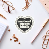 a heart-shaped sticker with the text "emotional support Kindle" surrounded by various office supplies such as a pen, a notebook, and a pair of glasses.