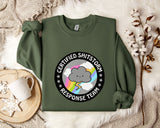 a green sweatshirt with a graphic design on it, placed on a bed with a white blanket and a mug.