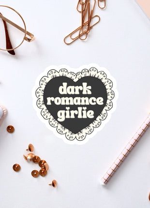 a heart-shaped sticker with the text "dark romance girlie" on a white background, surrounded by various office supplies such as a pen, a notebook, and a pair of glasses.