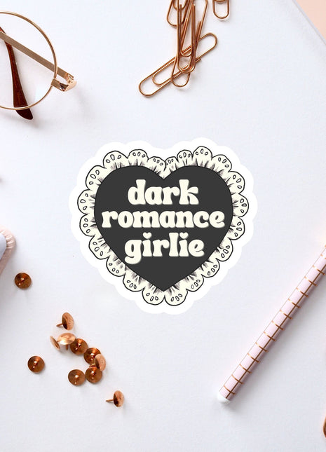 a heart-shaped sticker with the text "dark romance girlie" on a white background, surrounded by various office supplies such as a pen, a notebook, and a pair of glasses.