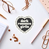 a heart-shaped sticker with the text "dark romance girlie" on a white background, surrounded by various office supplies such as a pen, a notebook, and a pair of glasses.