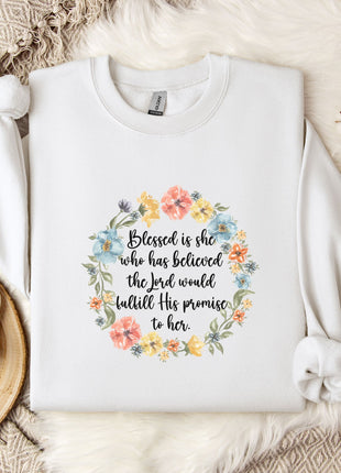 A white sweatshirt with a floral wreath design and a quote printed on it, placed on a bed with a white blanket and a white mug.