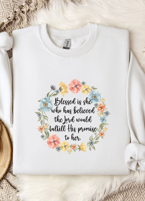 A white sweatshirt with a floral wreath design and a quote printed on it, placed on a bed with a white blanket and a white mug.