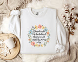 A white sweatshirt with a floral wreath design and a quote printed on it, placed on a bed with a white blanket and a white mug.