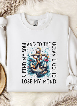 A white t-shirt with a graphic design of an anchor and waves, along with the text "I find my soul and to the ocean I go to lose my mind" printed on it. The t-shirt is laid out on a bed with a white blanket and a mug, and there are some flowers and a branch nearby.