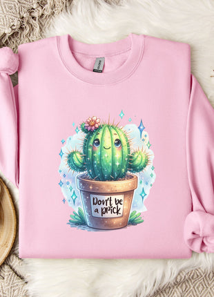 a pink sweatshirt with a cactus graphic and the text "Don't be a prick" printed on it. The sweatshirt is laid out on a bed with a white blanket and a small white mug. There are also some white flowers and a branch of cotton bolls in the background.
