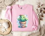 a pink sweatshirt with a cactus graphic and the text "Don't be a prick" printed on it. The sweatshirt is laid out on a bed with a white blanket and a small white mug. There are also some white flowers and a branch of cotton bolls in the background.