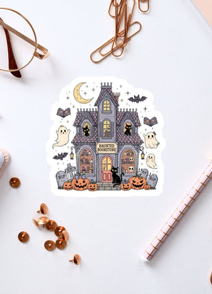 a Halloween-themed sticker of a house with bats, pumpkins, and ghostly figures, surrounded by various office supplies such as a pen, a notebook, and a pair of glasses.