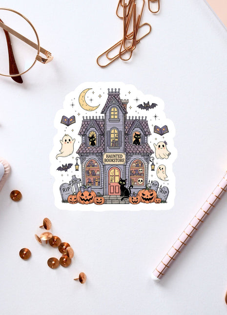 a Halloween-themed sticker of a house with bats, pumpkins, and ghostly figures, surrounded by various office supplies such as a pen, a notebook, and a pair of glasses.