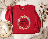 A red sweater with a floral design and a Bible verse printed on it, along with a mug and some white flowers on a white fur rug.