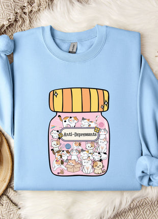 A light blue sweatshirt with a graphic of a jar containing various cartoon cats and the text "Anti-Depressants" printed on it, along with a mug and some flowers on a bed.