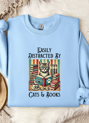 A light blue sweatshirt with a printed design of a cat surrounded by books and a cup of coffee, along with the text "Easily Distracted By Cats & Books".