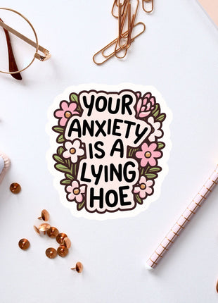 a sticker with the text "Your anxiety is a lying hoe" surrounded by various office supplies such as a pen, a pair of glasses, and a notebook.