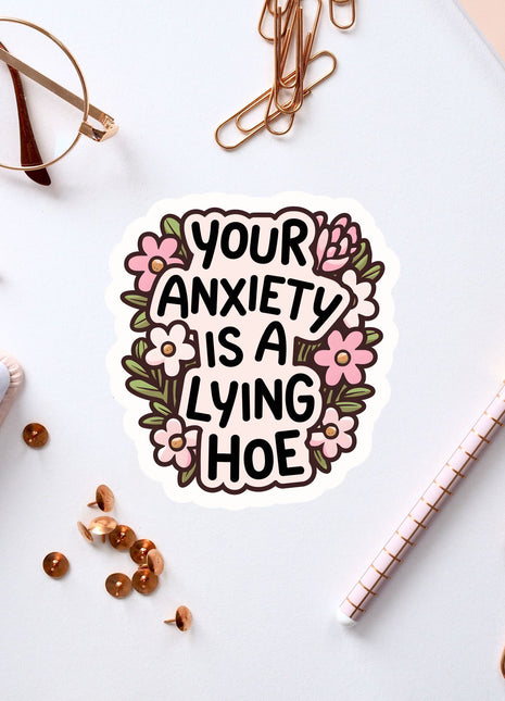 a sticker with the text "Your anxiety is a lying hoe" surrounded by various office supplies such as a pen, a pair of glasses, and a notebook.