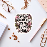 a sticker with the text "Your anxiety is a lying hoe" surrounded by various office supplies such as a pen, a pair of glasses, and a notebook.