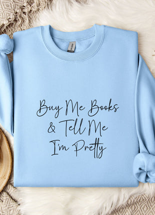 A light blue sweatshirt with the text "Buy Me Books & Tell Me I'm Pretty" printed on it, along with a mug and some flowers on a bed.