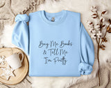 A light blue sweatshirt with the text "Buy Me Books & Tell Me I'm Pretty" printed on it, along with a mug and some flowers on a bed.
