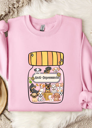 A pink sweatshirt with a graphic of a jar filled with cartoon dogs and a label that says "Anti-Depressants" is displayed on a bed with a white blanket and a small white mug.
