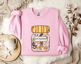 A pink sweatshirt with a graphic of a jar filled with cartoon dogs and a label that says "Anti-Depressants" is displayed on a bed with a white blanket and a small white mug.