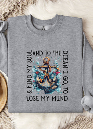 A gray sweatshirt with a graphic design of an anchor and waves, along with the text "AND I GO TO LOSE MY MIND" printed on it, is displayed on a bed with a white blanket and a mug.