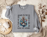 A gray sweatshirt with a graphic design of an anchor and waves, along with the text "AND I GO TO LOSE MY MIND" printed on it, is displayed on a bed with a white blanket and a mug.