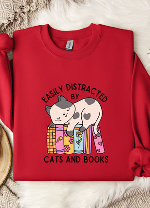 a red sweater with a cartoon cat and the text "EASILY DISTracted BY CATS AND BOOKS" printed on it, along with a mug and a plant.