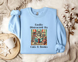 A light blue sweatshirt with a printed design of a cat reading a book, accompanied by the text "Easily Distracted By Cats & Books".