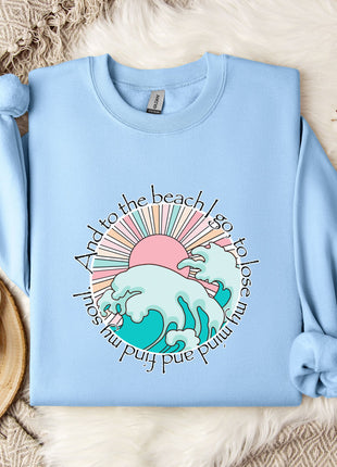 A light blue sweatshirt with a graphic design of a wave and sun, along with the text "And to the beach I go, to lose sight of you while I'm busy and busy". The sweatshirt is laid out on a bed with a white blanket and a mug, and there are some cotton balls and a snowflake decoration nearby.