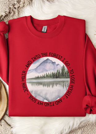 A red sweater with a circular design featuring a mountain landscape and a lake, along with the text "John Muir - AND INTO THE FOREST I GO TO LOSE MY SELF" printed on it. The sweater is laid out on a white fur rug, with a white mug, a snowflake, and a branch of cotton bolls nearby.