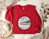 A red sweater with a circular design featuring a mountain landscape and a lake, along with the text "John Muir - AND INTO THE FOREST I GO TO LOSE MY SELF" printed on it. The sweater is laid out on a white fur rug, with a white mug, a snowflake, and a branch of cotton bolls nearby.