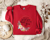 a red sweater with a floral design and text printed on it, placed on a white fur rug. There is also a white mug and a branch of cotton flowers nearby.