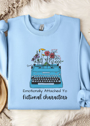 A light blue sweatshirt with a graphic of a typewriter filled with flowers, accompanied by the text "Emotionally Attached To Fictional Characters".