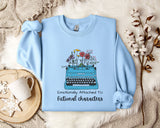 A light blue sweatshirt with a graphic of a typewriter filled with flowers, accompanied by the text "Emotionally Attached To Fictional Characters".