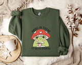 A green sweatshirt with a mushroom graphic and the text "HANG IN THERE IT GETS WORSE" printed on it, along with a mug and some white flowers on a bed.