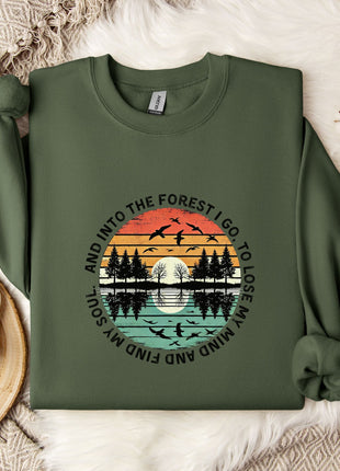 A green sweatshirt with a graphic design depicting a sunset over a forest, along with the text "And into the forest go I lose my mind" printed on it. The sweatshirt is laid out on a bed with a white blanket and a white mug with a snowflake design.