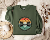 A green sweatshirt with a graphic design depicting a sunset over a forest, along with the text "And into the forest go I lose my mind" printed on it. The sweatshirt is laid out on a bed with a white blanket and a white mug with a snowflake design.
