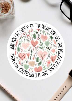 a round sticker with a floral design and text, surrounded by various office supplies such as a pair of glasses, a watch, and a pen.
