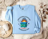A light blue sweatshirt with a graphic design depicting a trash can on fire, surrounded by flames and smoke, along with the text "Emotional Support Dumpster Fire".