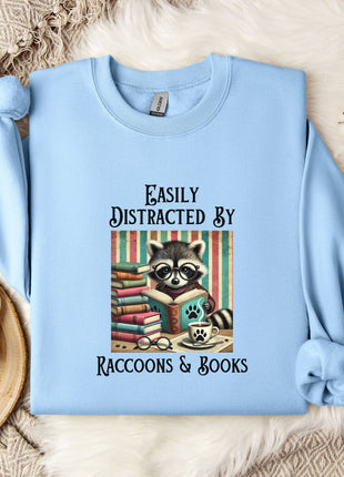 A light blue sweatshirt with a graphic design of a raccoon reading books, accompanied by the text "Easily Distracted By Raccoons & Books".