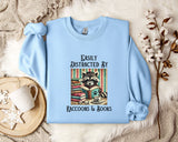 A light blue sweatshirt with a graphic design of a raccoon reading books, accompanied by the text "Easily Distracted By Raccoons & Books".