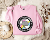 a pink sweatshirt with a graphic design on it, placed on a bed with a white blanket and a mug.