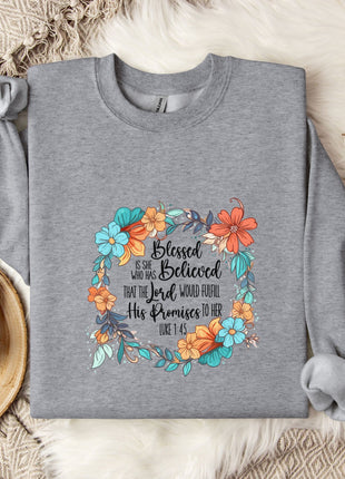 A gray sweatshirt with a floral design and text printed on it, along with a mug, a branch of cotton flowers, and a blanket on a bed.