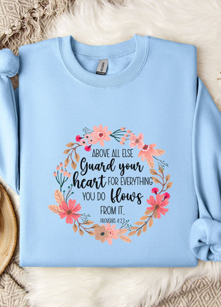 A light blue sweatshirt with a floral design and text printed on it, placed on a bed with a white blanket and a white mug.