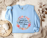 A light blue sweatshirt with a floral design and text printed on it, placed on a bed with a white blanket and a white mug.